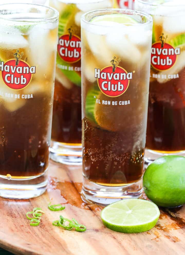 Four tall Havana Club glasses with Cuba Libre cocktails garnished with lime on a wood tray.