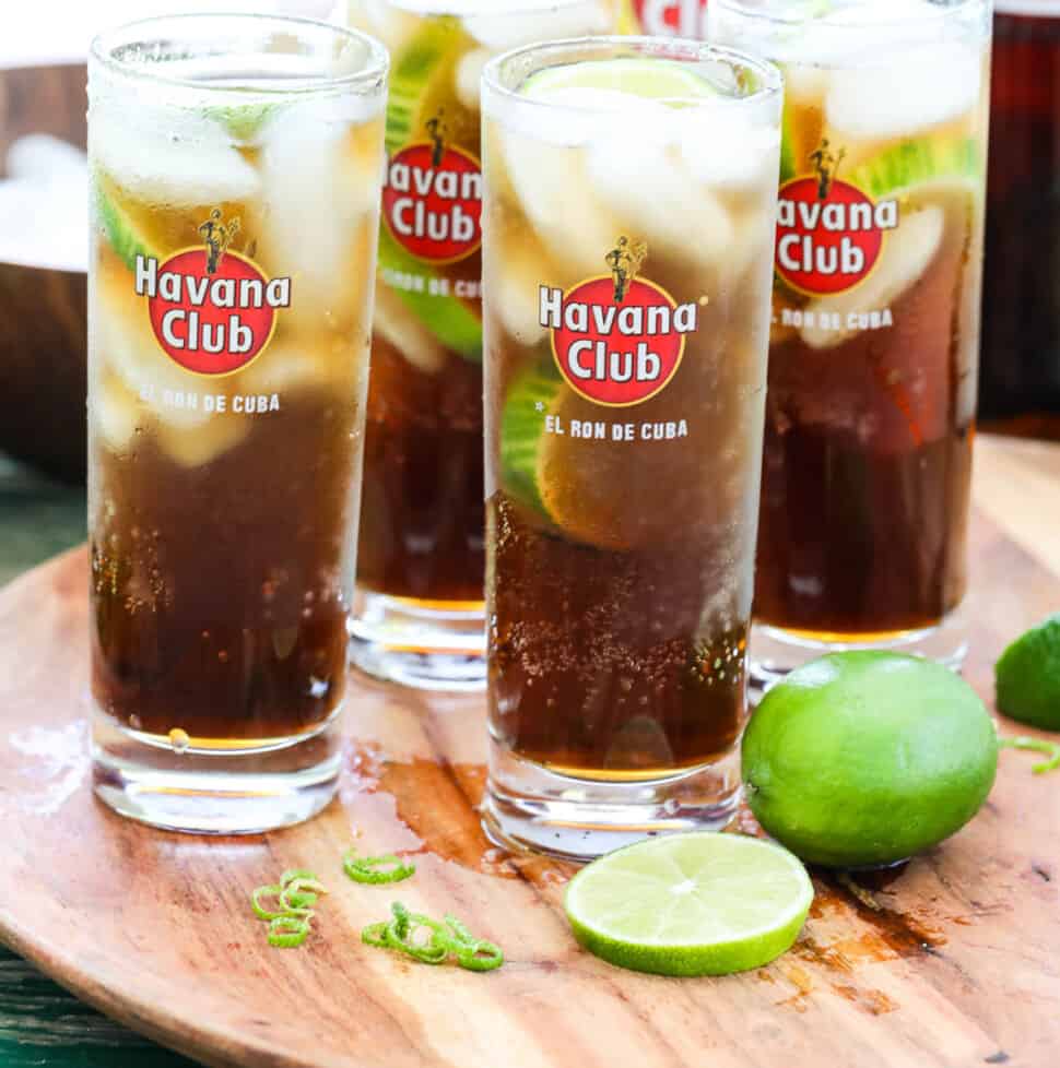 Four tall Havana Club glasses with Cuba Libre cocktails garnished with lime on a wood tray.