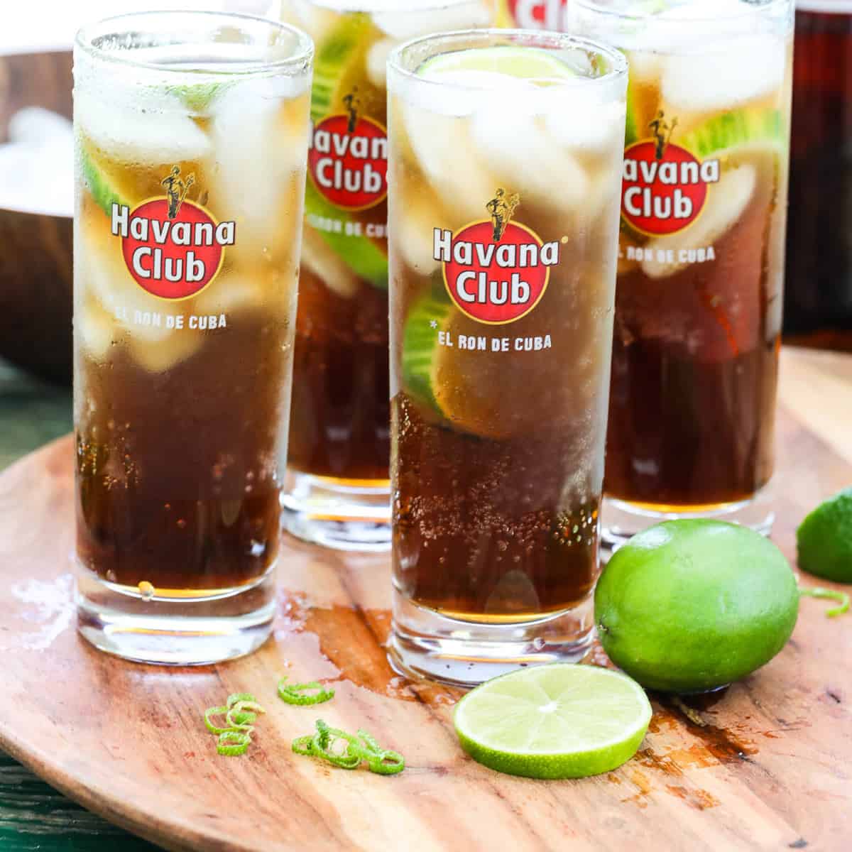 Four tall Havana Club glasses with Cuba Libre cocktails garnished with lime on a wood tray.