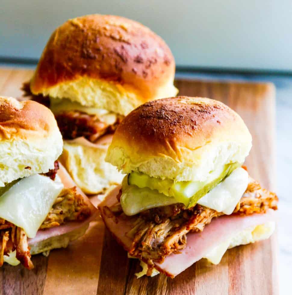 Three pulled pork sandwiches on Hawaiian buns with pickles, cheese and ham on a cutting board.