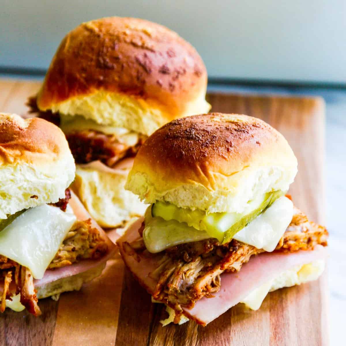 Three pulled pork sandwiches on Hawaiian buns with pickles, cheese and ham on a cutting board.