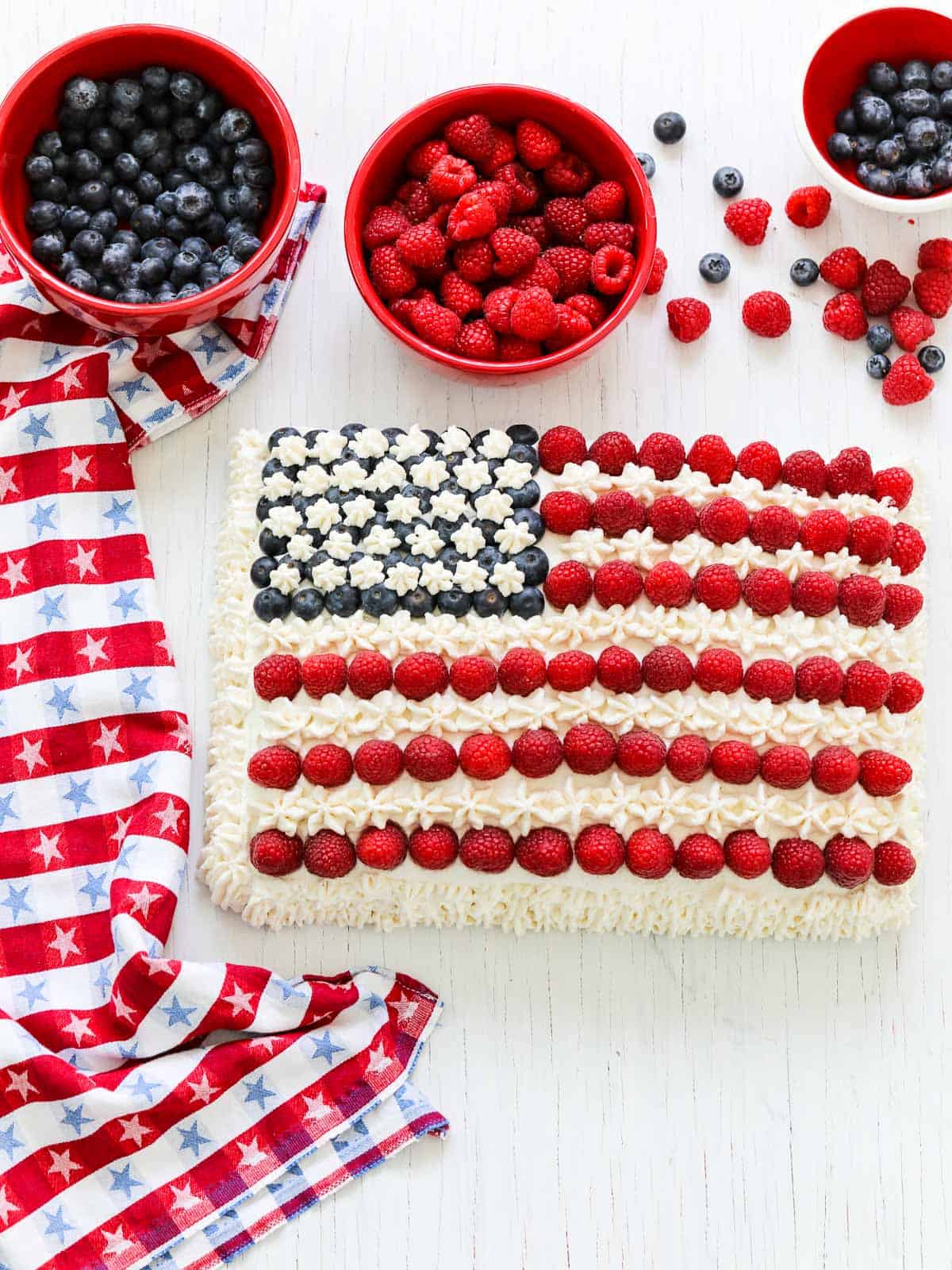 An American flag cake decorated with blueberries, raspberries, and white frosting with a patriotic star towel.