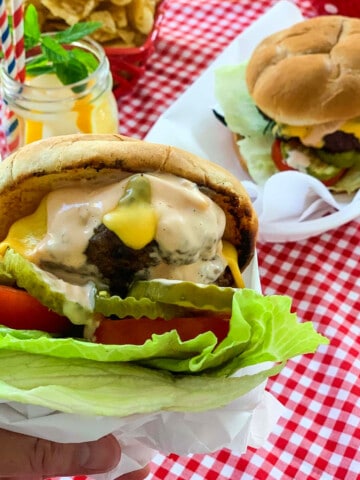 A person holding a messy grilled burger topped with melted cheese, burger sauce, lettuce, tomato, and pickles.