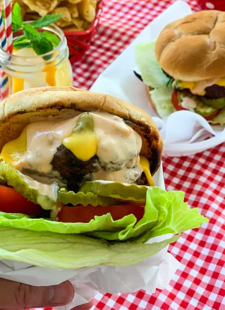 A person holding a messy grilled burger topped with melted cheese, burger sauce, lettuce, tomato, and pickles.