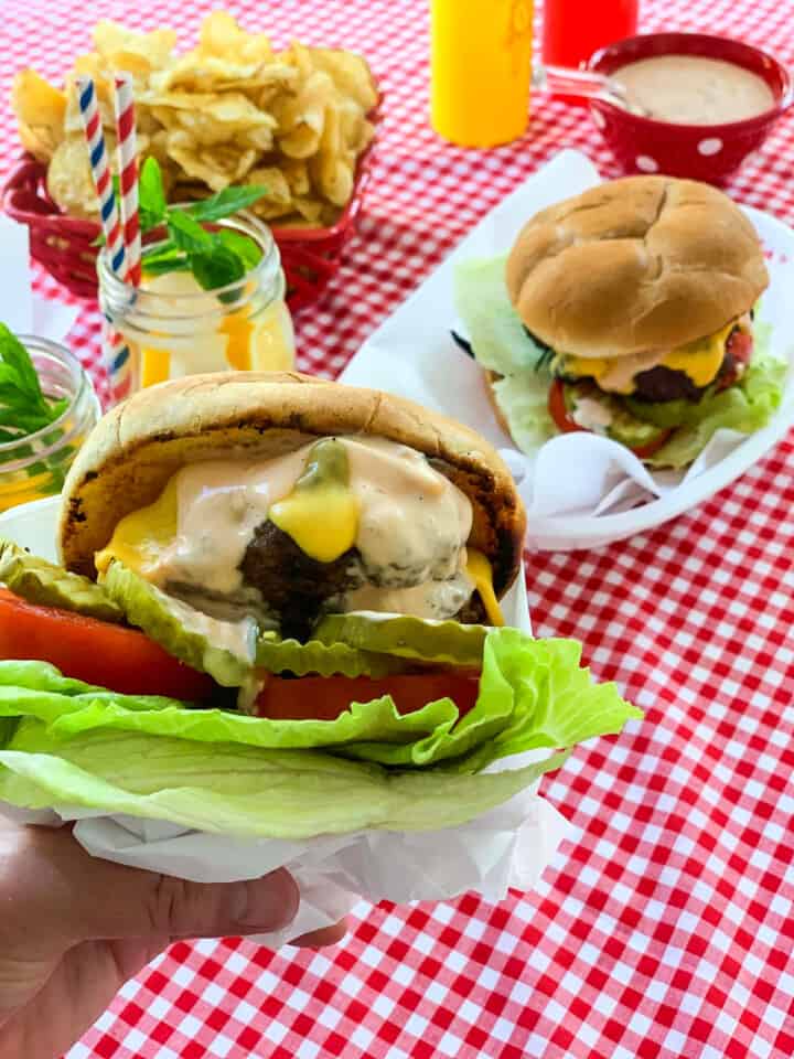 A lady holding a messy grilled hamburger wrapped in paper with baskets of burgers in the background.