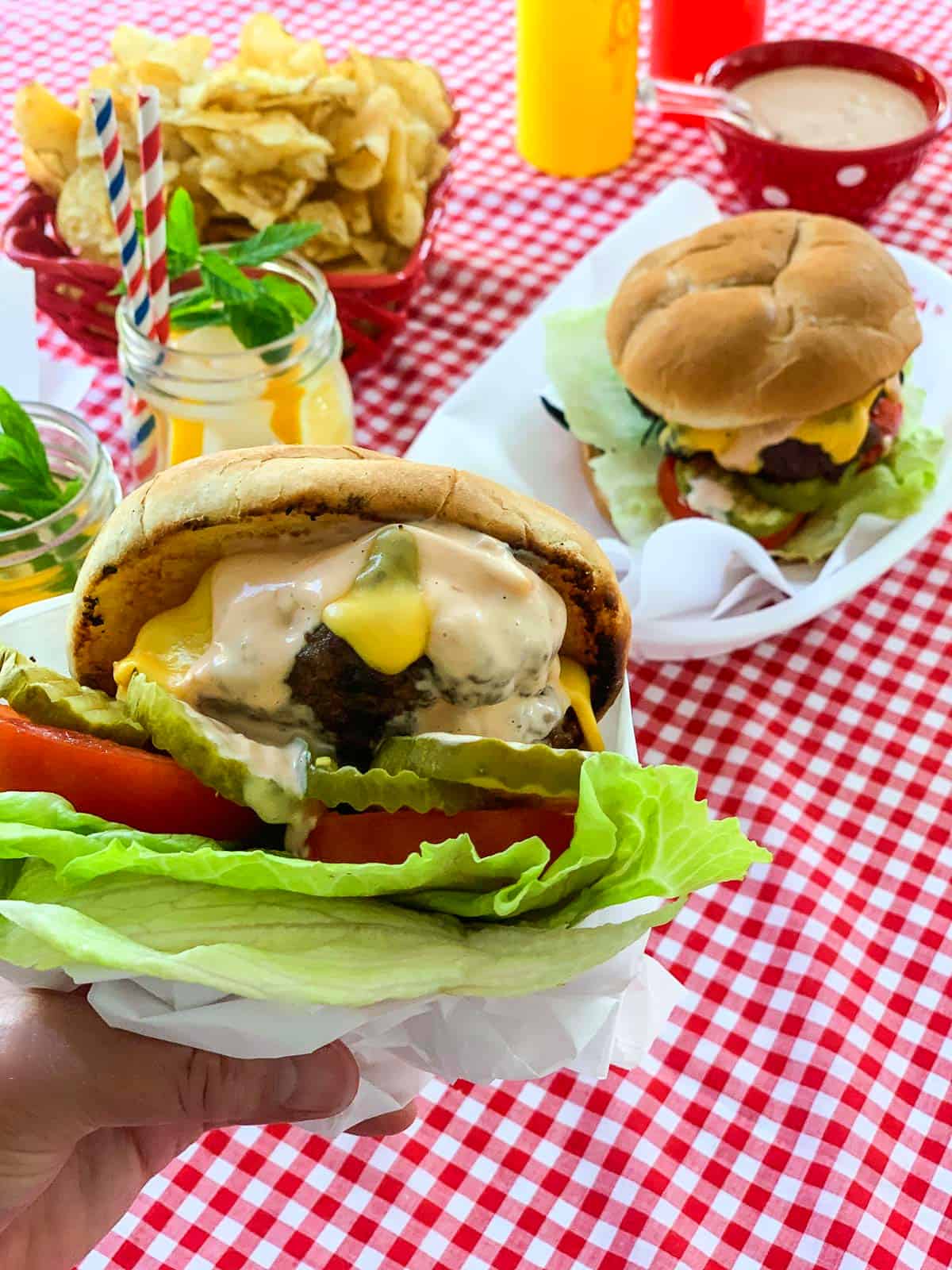 A lady holding a messy grilled cheeseburger wrapped in paper with baskets of burgers in the background.