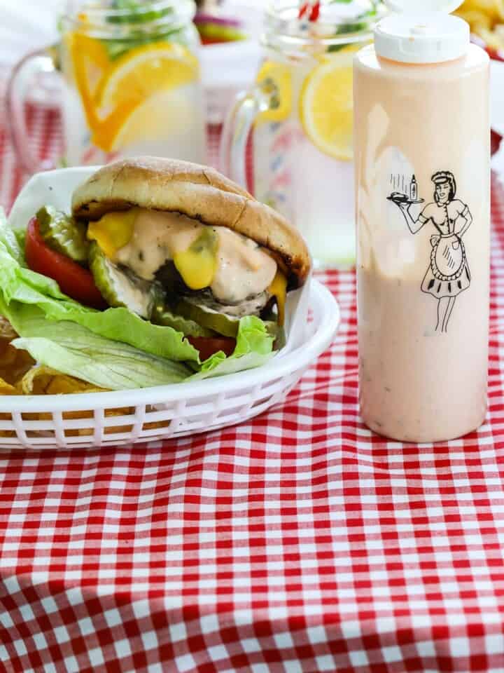 A messy grilled burger topped with lettuce, tomato, cheese, pickles and burger sauce in a basket with a bottle of burger sauce.