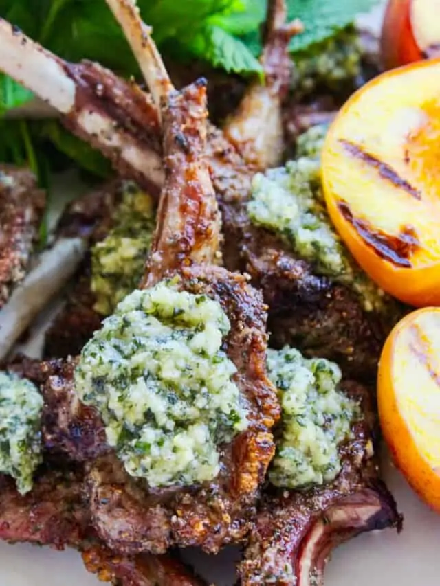 A white platter filled with grilled lamb chops topped with mint pesto and grilled peaches.