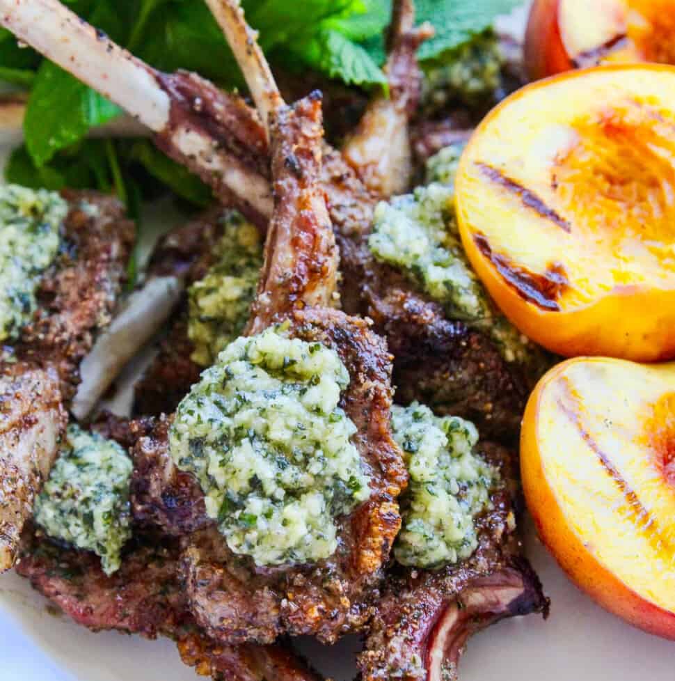 A white platter filled with grilled lamb chops topped with mint pesto and grilled peaches.