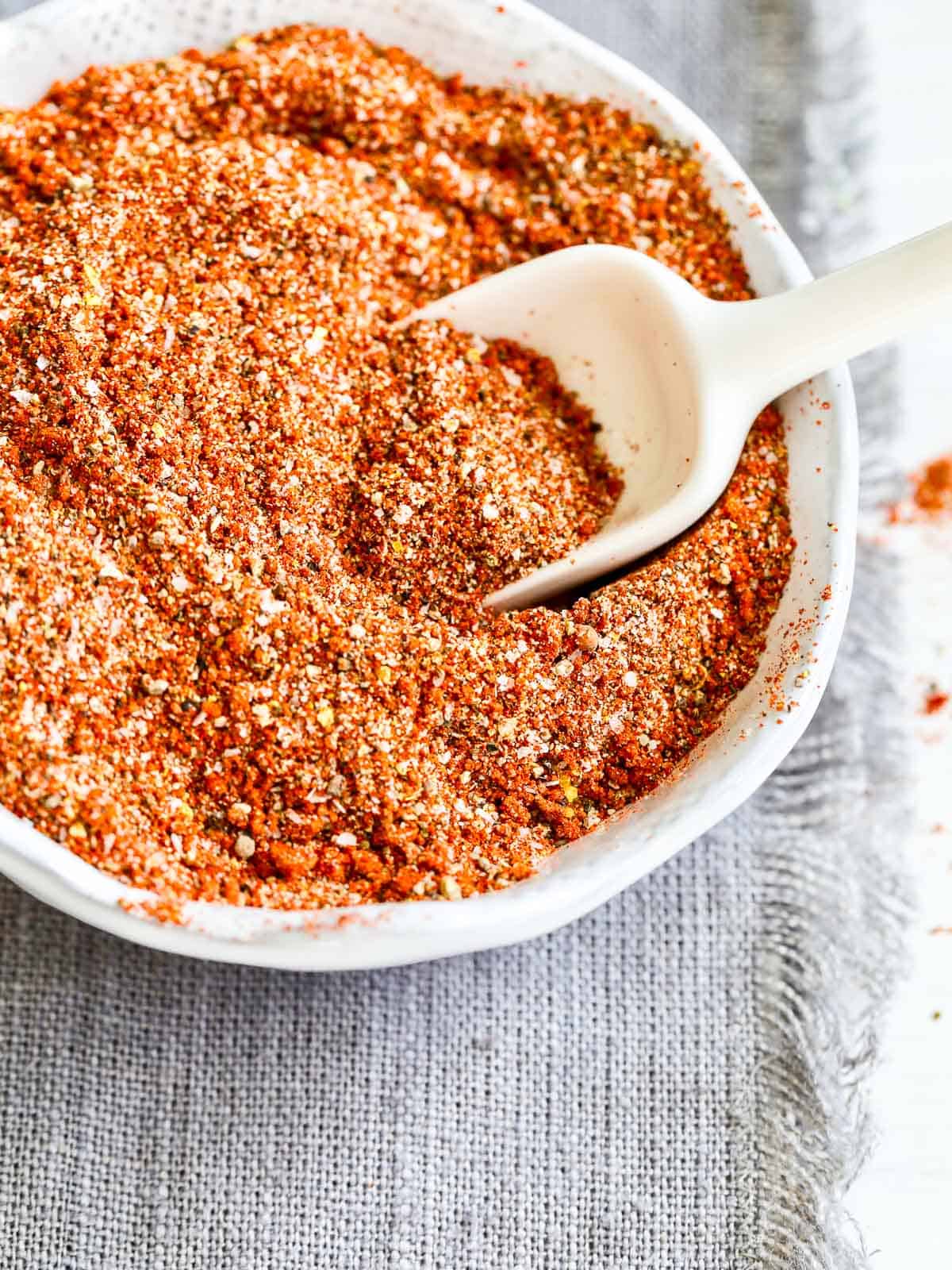 A small white bowl filled with a dry rub for BBQ and grilling with a small white scoop stuck in the seasoning.