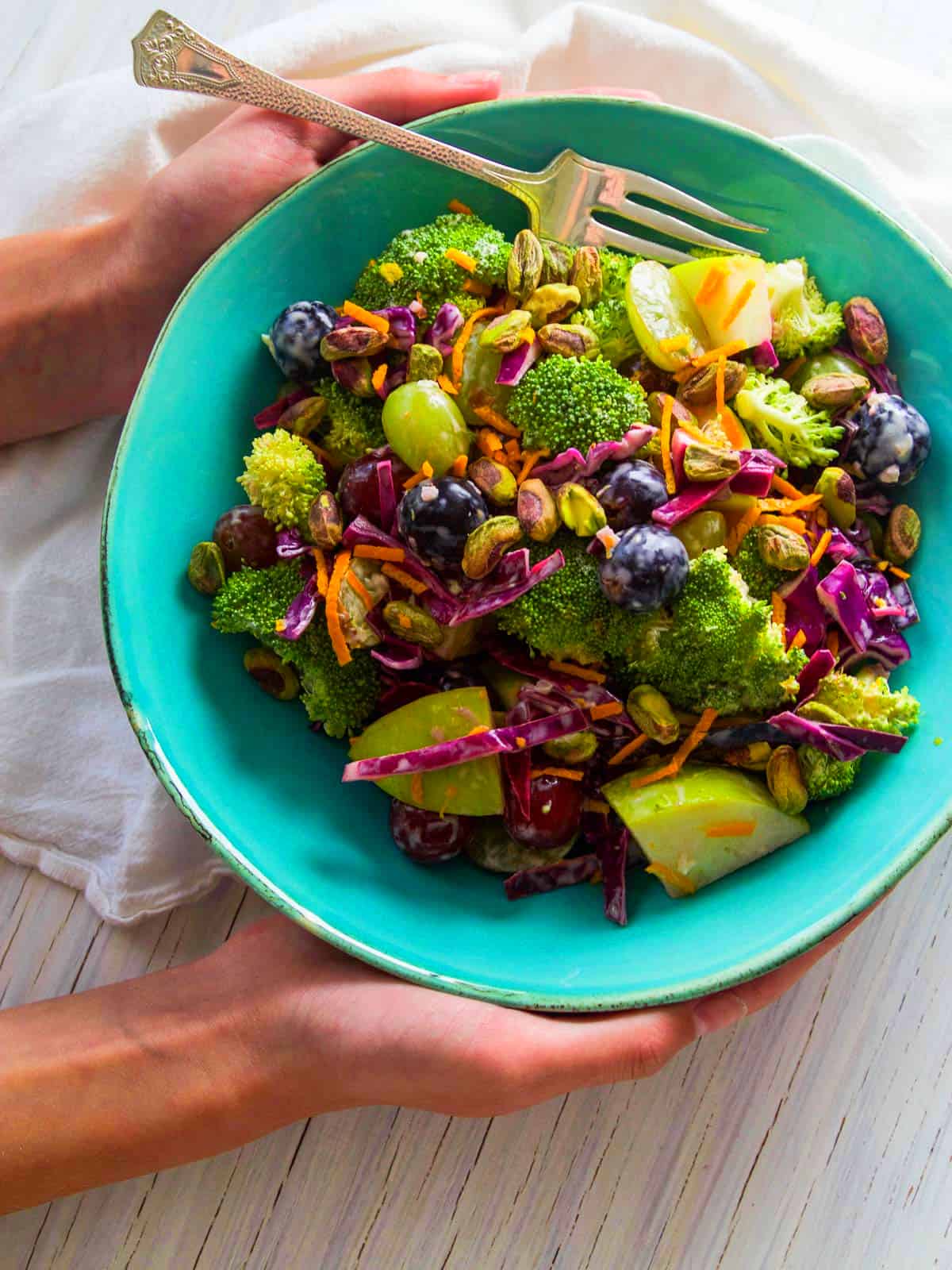 A young lady holding a turquoise bowl filled with broccoli salad drizzled with orange dressing and pistachios.