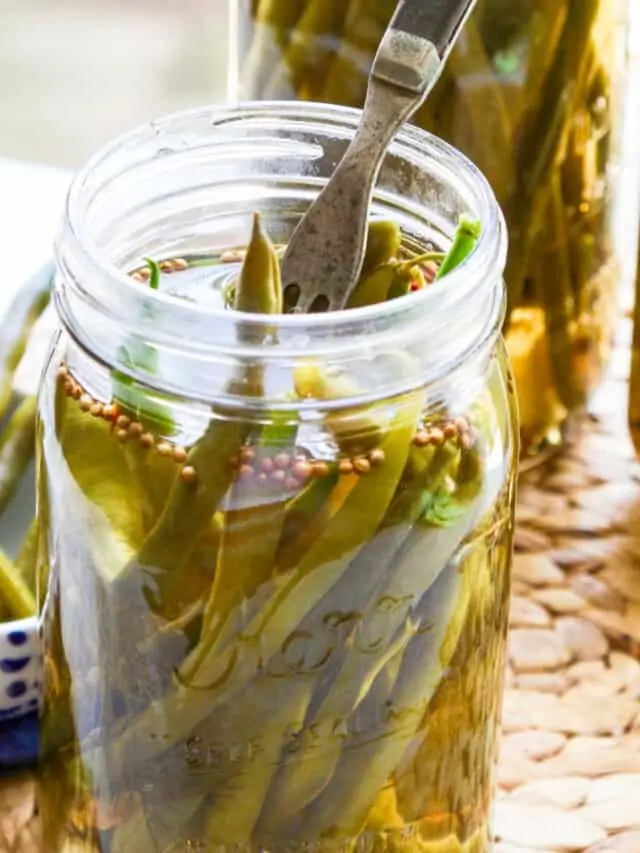 Three jars of pickled green beans with a fork stuck inside the jar and some in a dish.