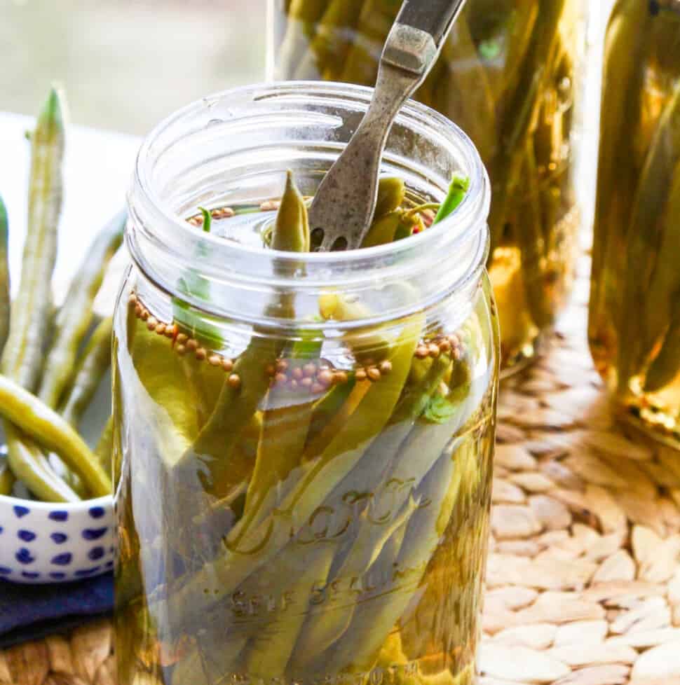 Three jars of pickled green beans with a fork stuck inside the jar and some in a dish.
