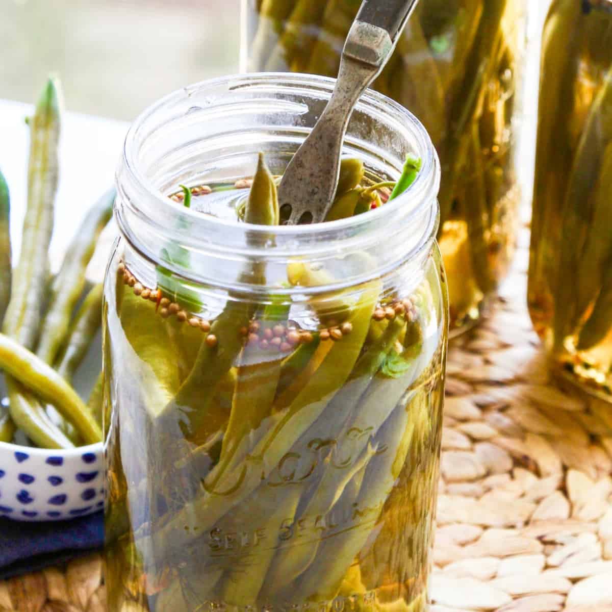 Three jars of pickled green beans with a fork stuck inside the jar and some in a dish.