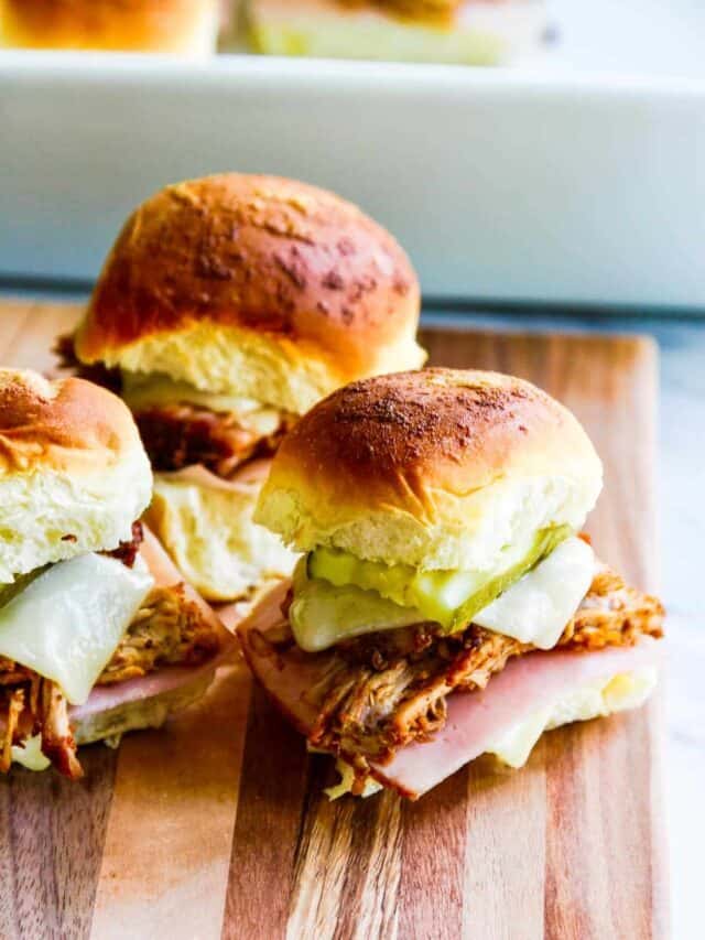 Three Cuban Pulled Pork sliders on a wooden board loaded with cheese and pickles.