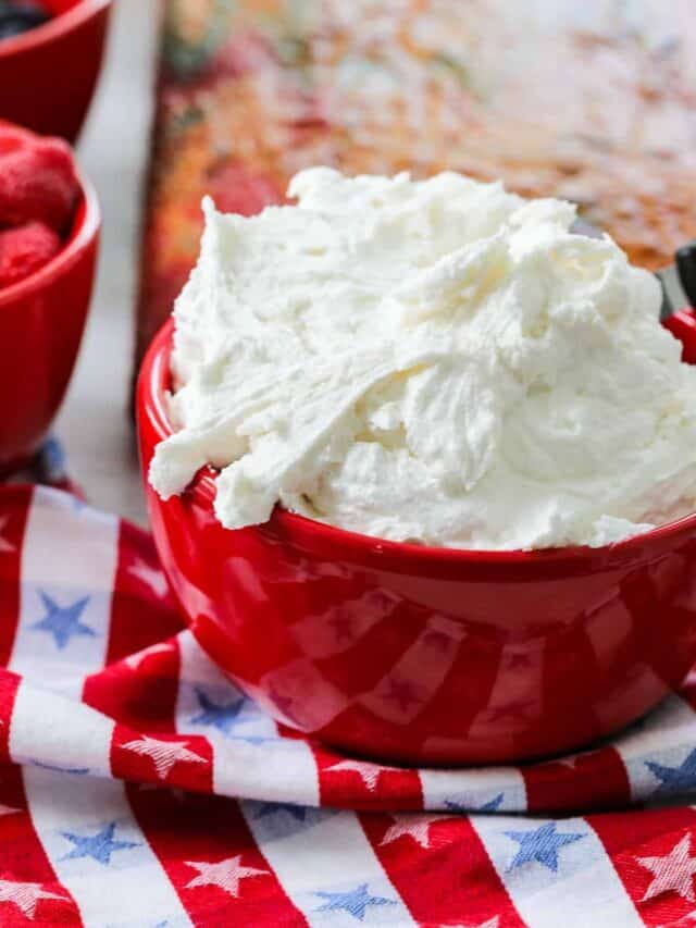 A red bowl filled with fluffy white buttercream frosting on a red white and blue stars and stripe towel.