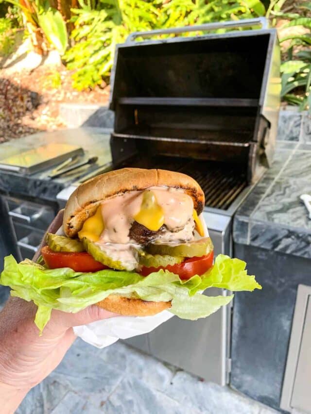 A lady holding a messy hamburger wrapped in parchment paper out by an open stainless steel gas grill.