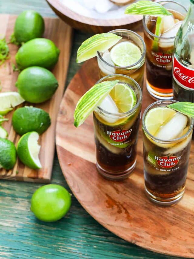 A wood tray filled with four Cuba Libre cocktails with limes slice on a cutting board.