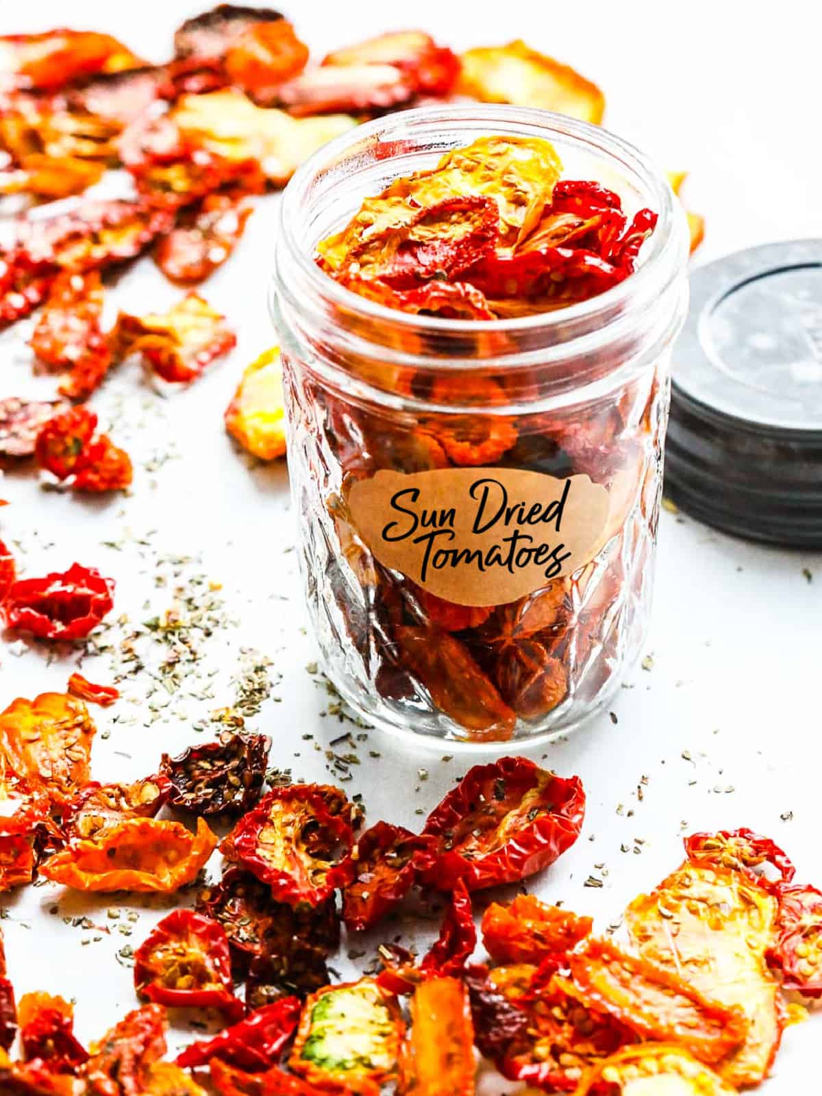 A small clear jar holding sun dried tomatoes with a label and them sprinkled on a table.