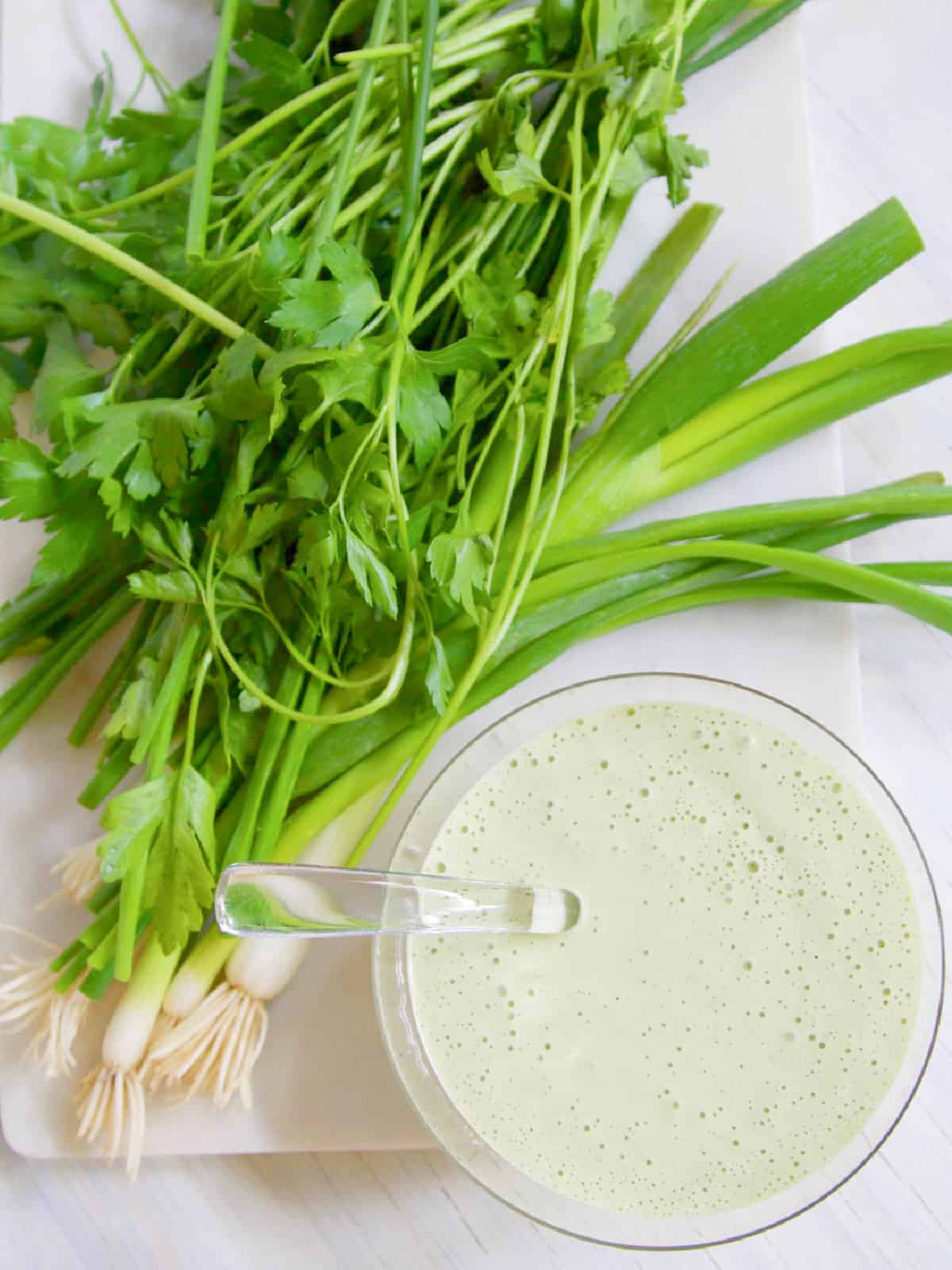 Clear glass bowl with glass spoon and a pile of green onions and parsley. 