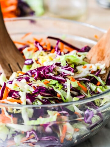 A clear glass bowl filled with tri color cole slaw and wooden tongs stuck in the slaw.