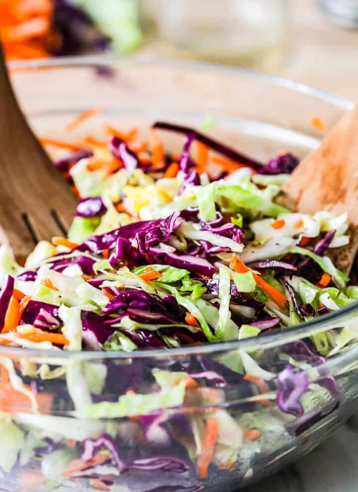 A clear glass bowl filled with tri color cole slaw and wooden tongs stuck in the slaw.