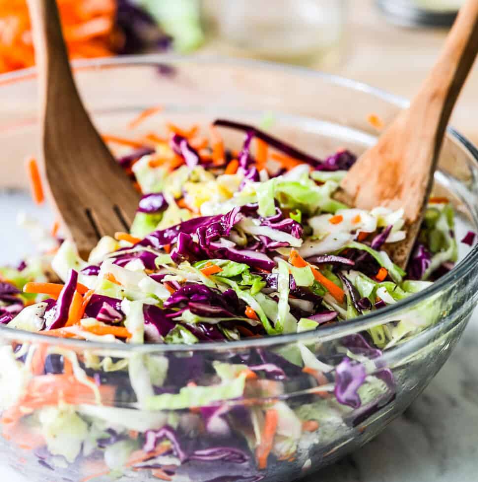 A clear glass bowl filled with tri color cole slaw and wooden tongs stuck in the slaw.