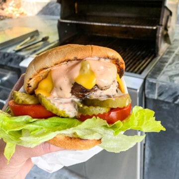 A lady holding a messy burger out by a gas grill topped with melted cheese, pickle chips, tomato, and wrapped in paper.