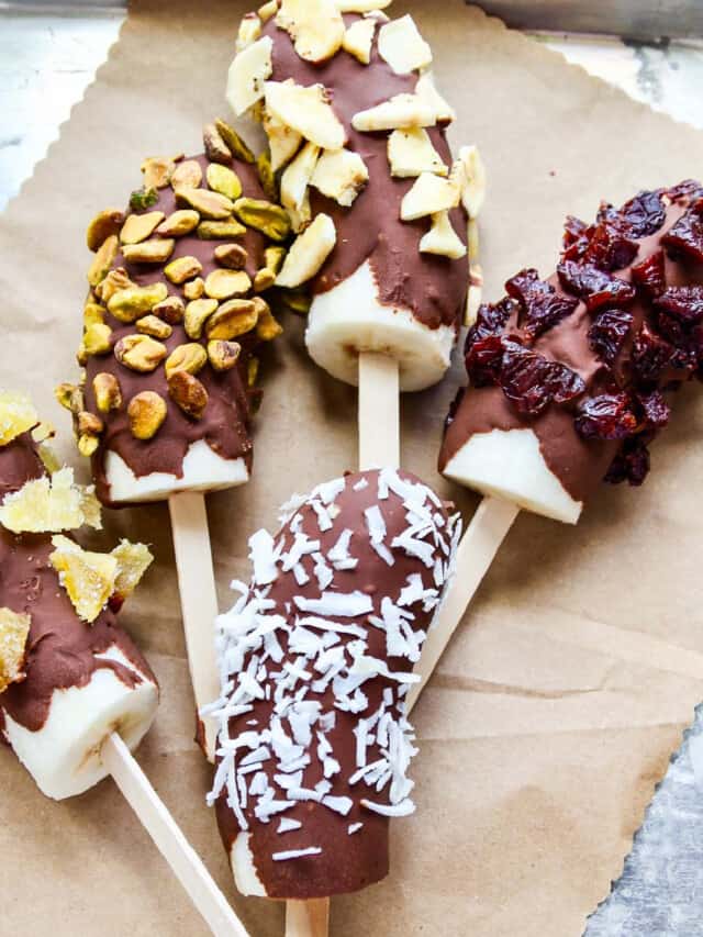 A small aluminum sheet pan with brown paper and 5 frozen bananas chocolate dipped and coated in nuts and dried fruit.