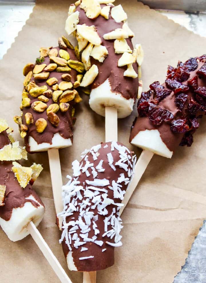 A small aluminum sheet pan with brown paper and 5 frozen bananas chocolate dipped and coated in nuts and dried fruit.