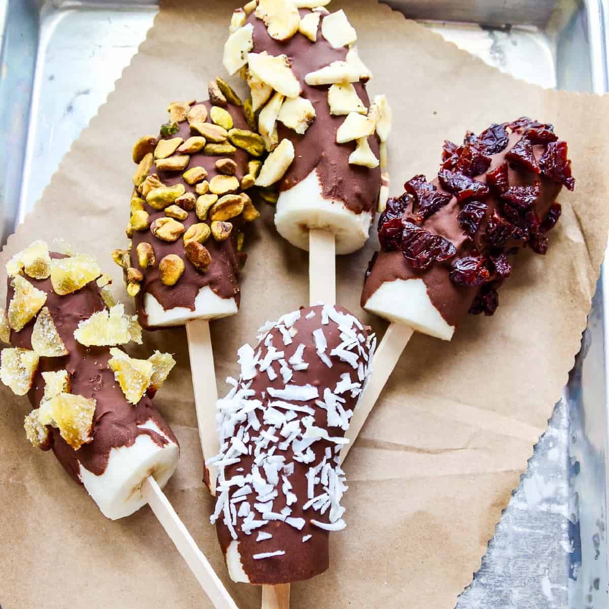A small aluminum sheet pan with brown paper and 5 frozen bananas chocolate dipped and coated in nuts and dried fruit.