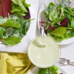 A small glass bowl with green goddess dressing and white plates of salad on a table.