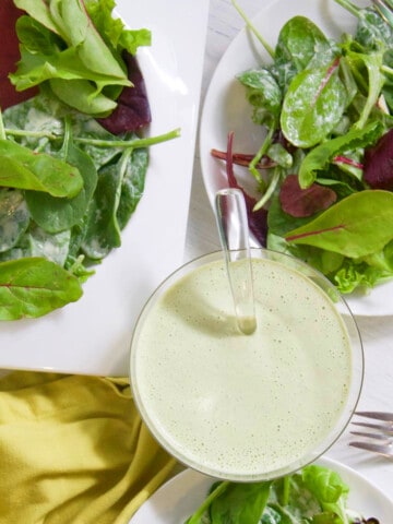 A small glass bowl with green goddess dressing and white plates of mixed salad greens on a table.