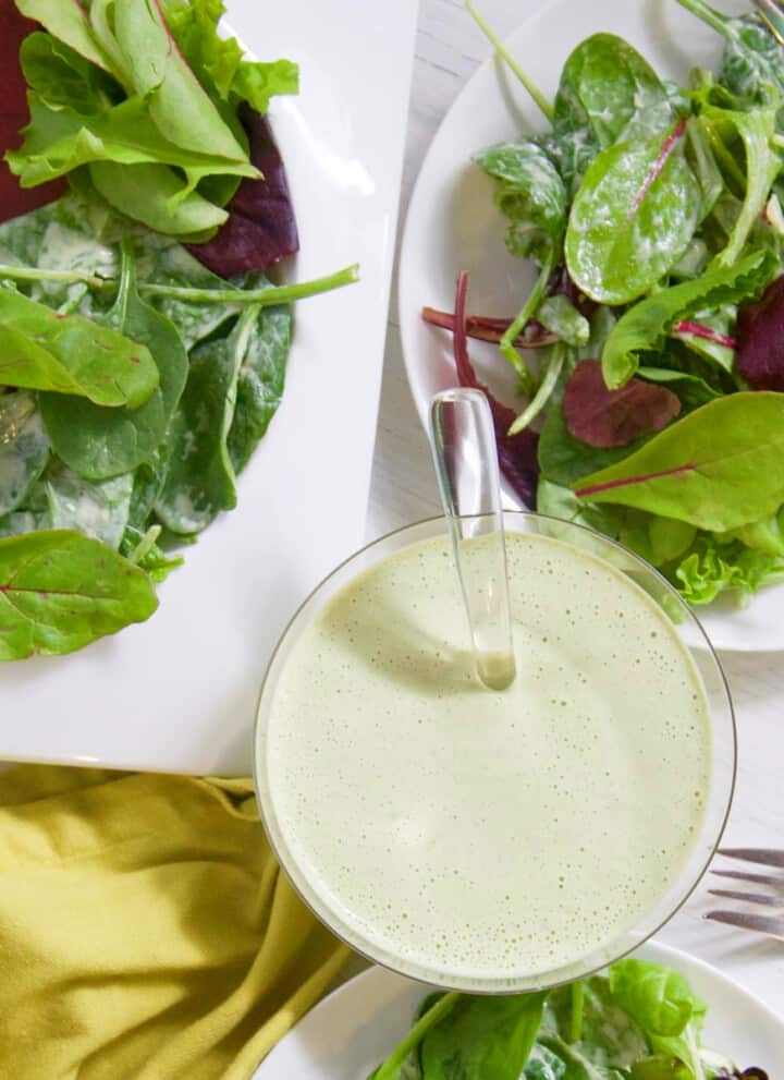 A small glass bowl with green goddess dressing and white plates of mixed salad greens on a table.