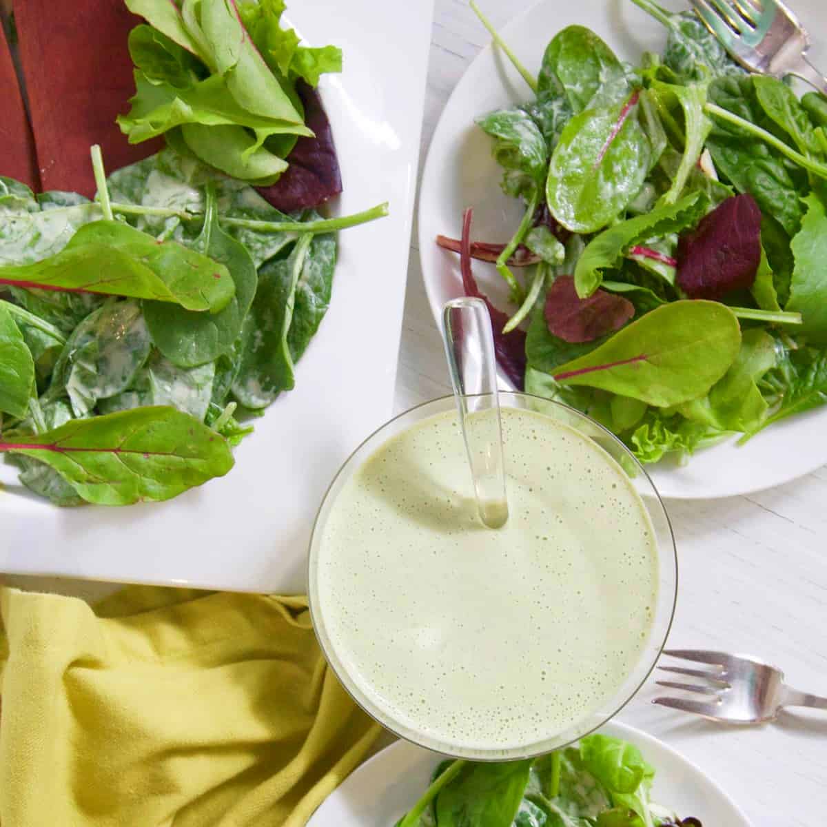 A small glass bowl with green goddess dressing and white plates of mixed salad greens on a table.