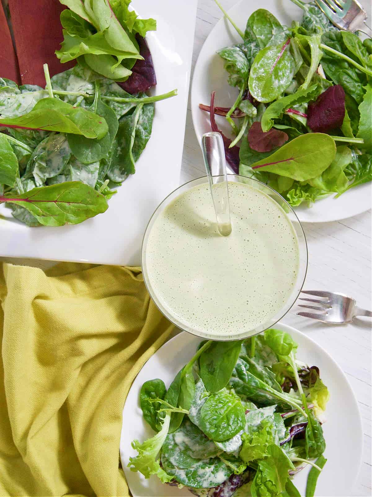 Two white plates with salad green tossed in green goddess dressing and  silver forks. 