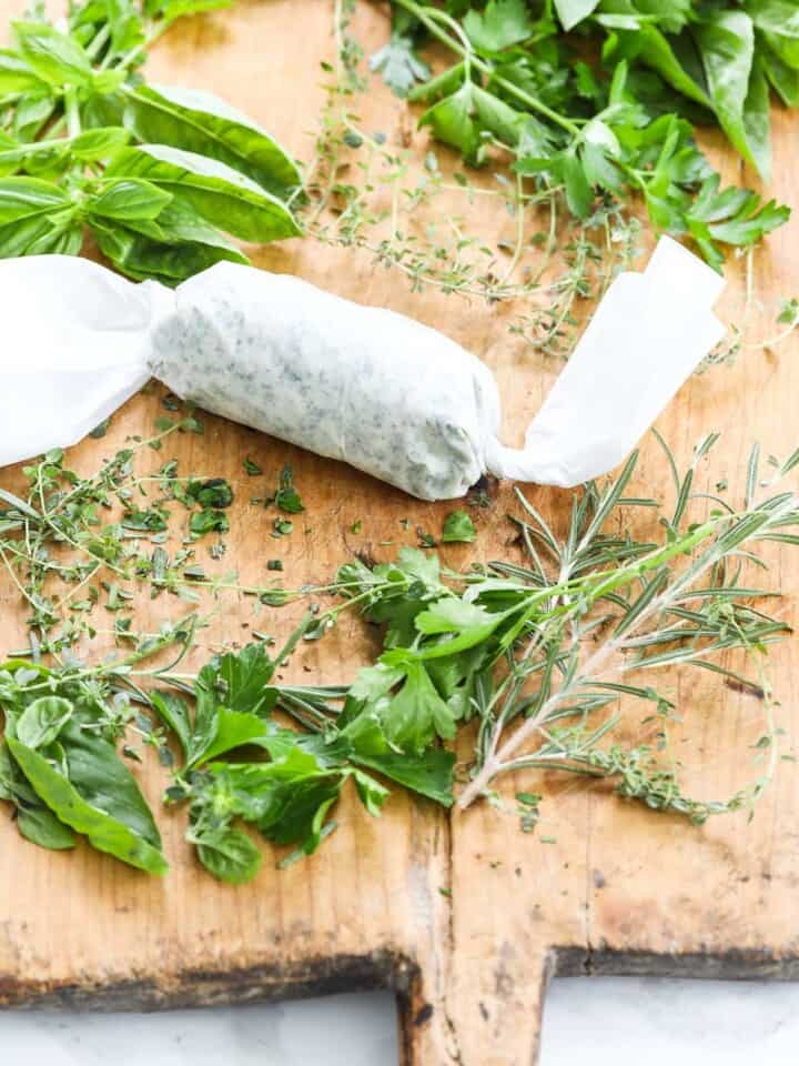 Herbed butter wrapped in parchment paper on a vintage cutting board with fresh herbs.