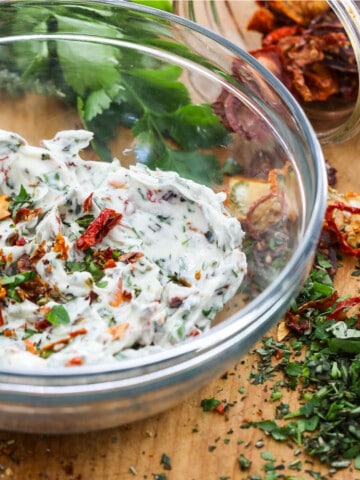 A glass bowl filled with compound butter made with herbs and sun dried tomatoes, and them scattered on the cutting board.