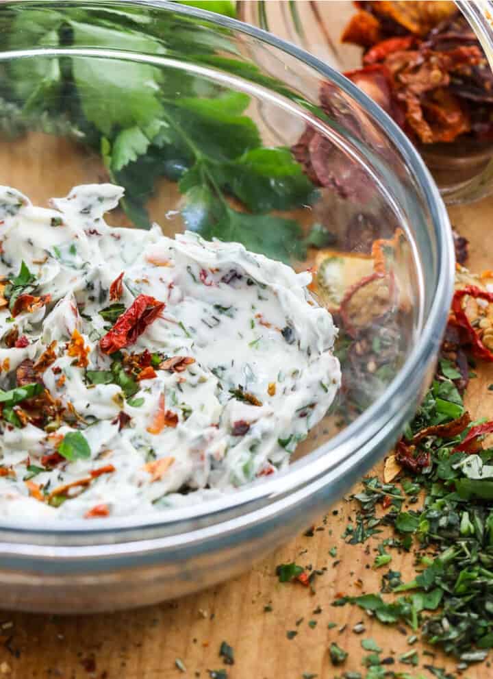 A glass bowl filled with compound butter made with herbs and sun dried tomatoes, and them scattered on the cutting board.