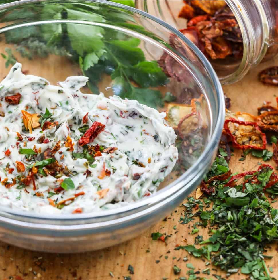 A glass bowl filled with compound butter made with herbs and sun dried tomatoes, and them scattered on the cutting board.