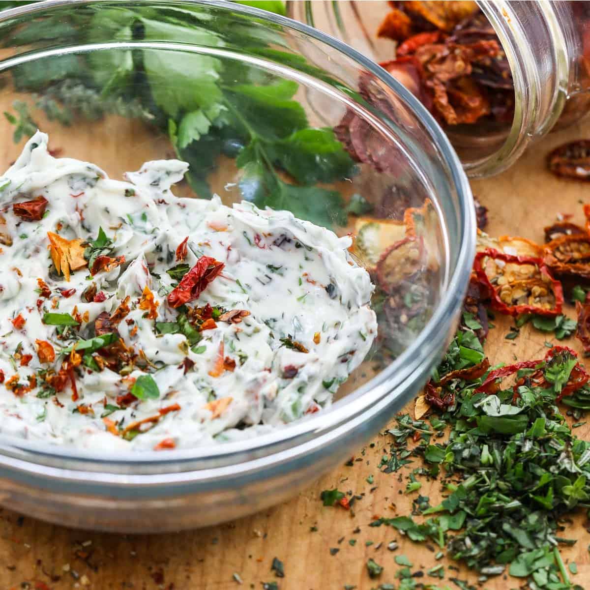 A glass bowl filled with compound butter made with herbs and sun dried tomatoes, and them scattered on the cutting board.