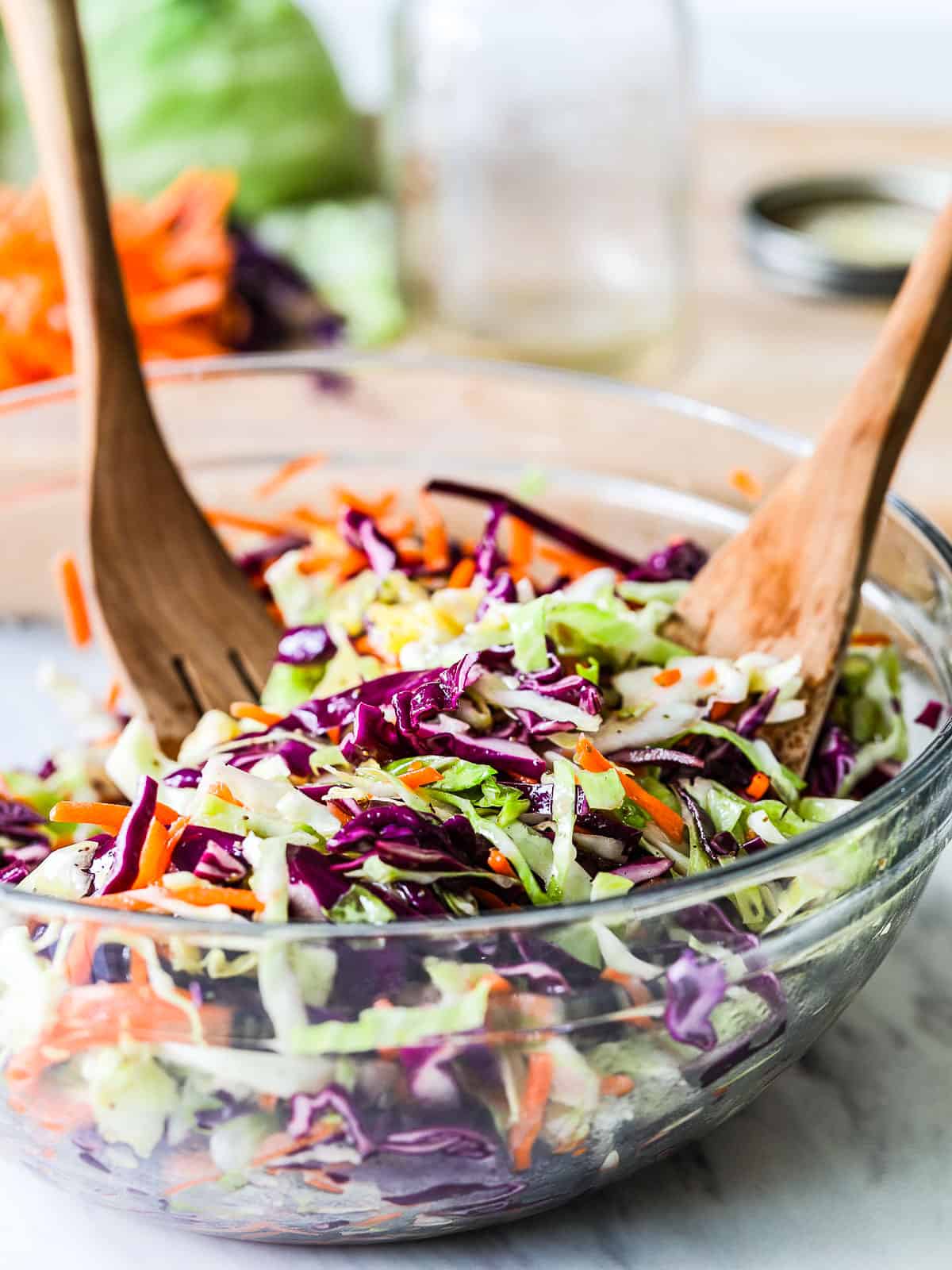 A large clear bowl filled with colorful cole slaw made with purple and green cabbage and carrots with wood serving tongs.
