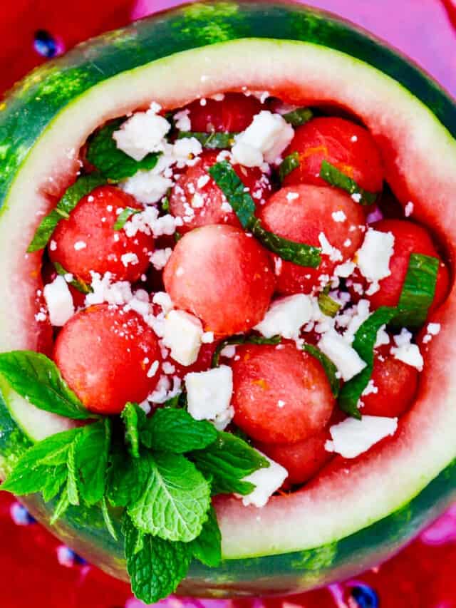 Looking down into a scooped out watermelon filled with rounded watermelon balls garnished with mint and feta cheese.