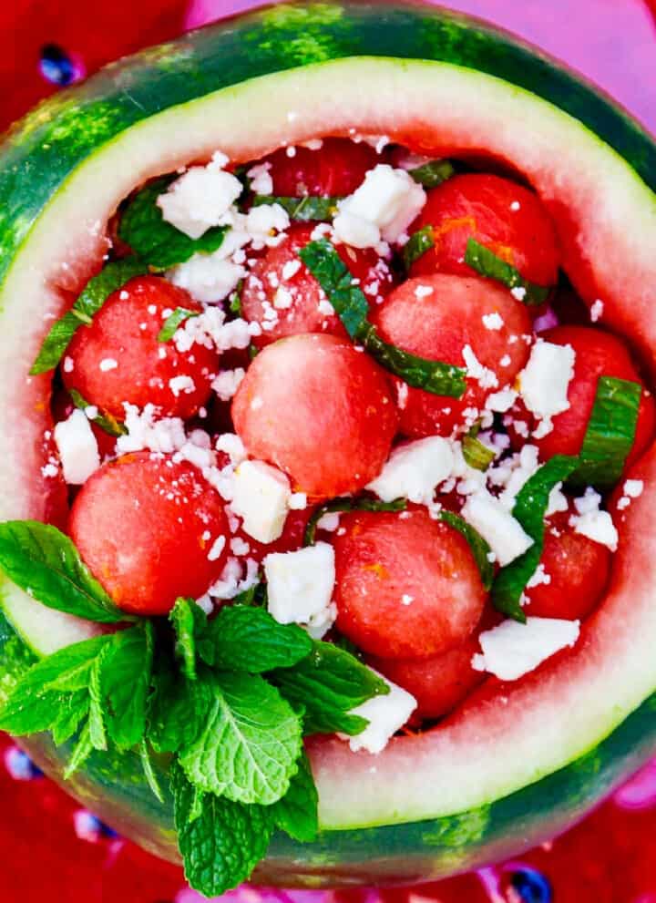 Looking down into a scooped out watermelon filled with rounded watermelon balls garnished with mint and feta cheese.
