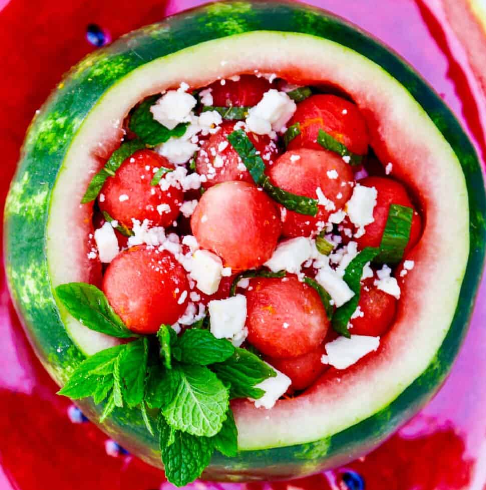 Looking down into a scooped out watermelon filled with rounded watermelon balls garnished with mint and feta cheese.