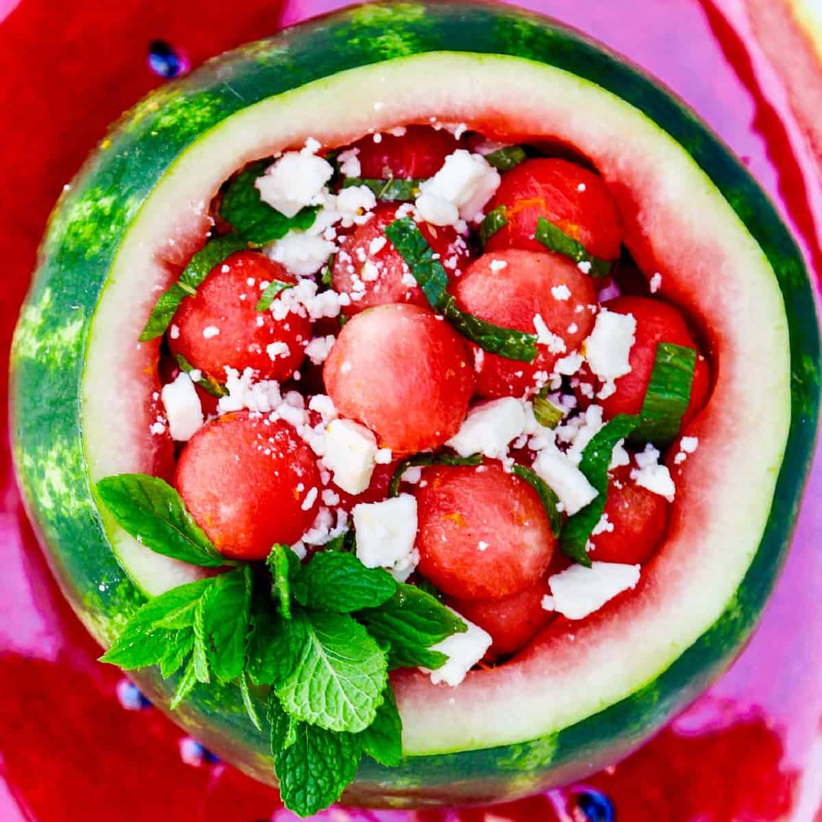 Looking down into a scooped out watermelon filled with rounded watermelon balls garnished with mint and feta cheese.