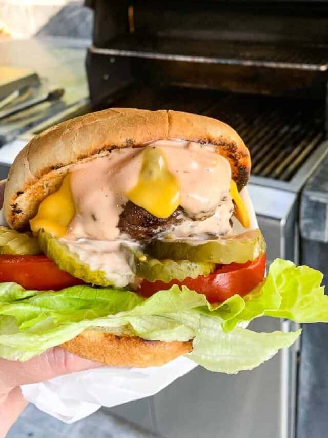 A lady holding a cheeseburger with lettuce, tomato, pickles, cheese, and burger sauce by an open grill.