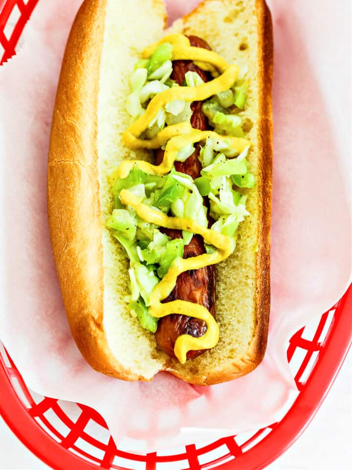 A hot dog with homemade relish and mustard toppings in a red plastic basket lined with paper.