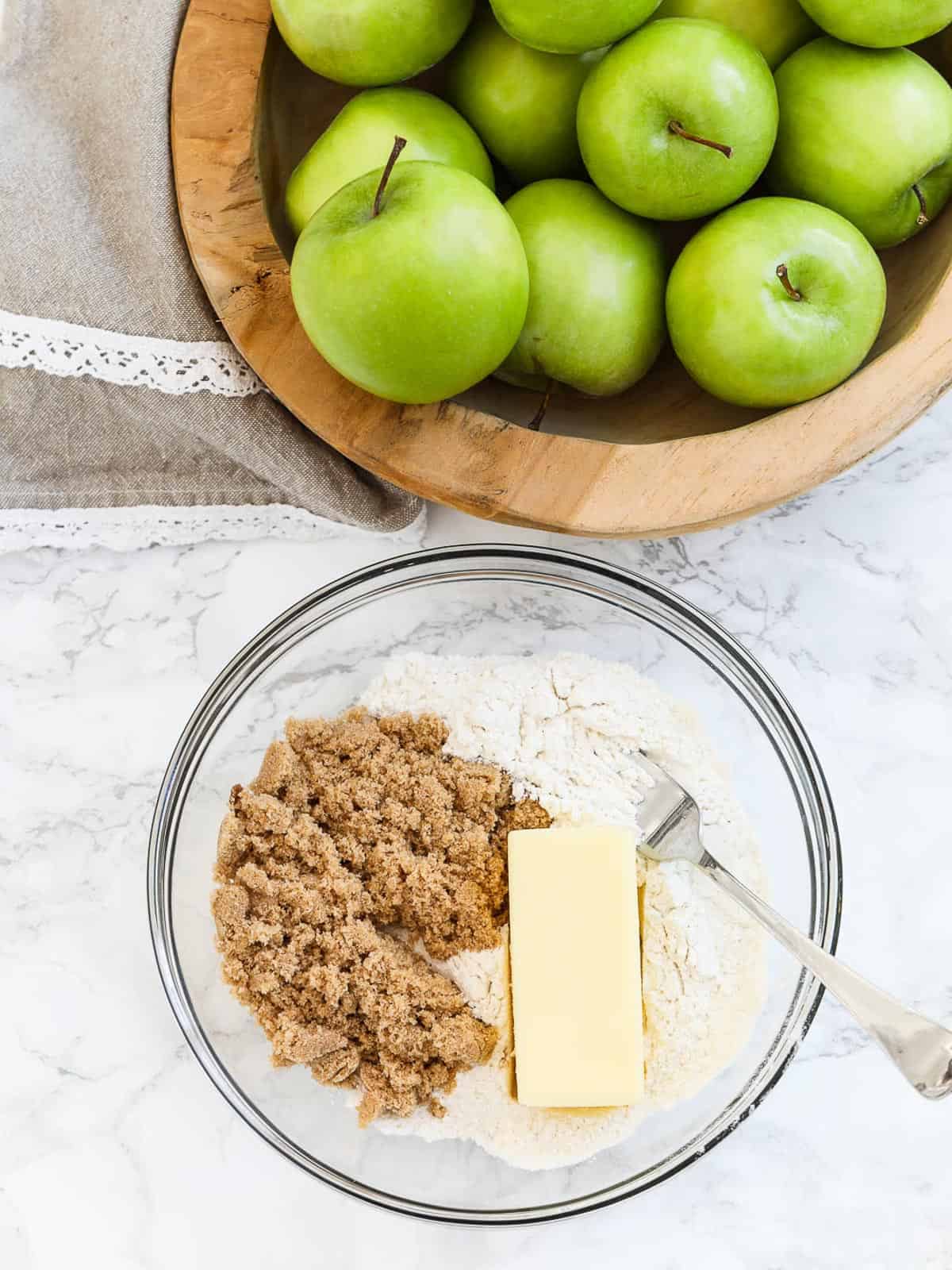 A clear glass bowl with flour, brown sugar, and a cube of butter with a fork to make crumbled topping for apple crumble pie.