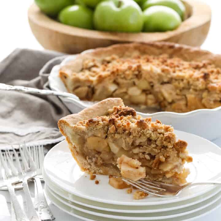 A white pie dish with a slice of apple crumble pie, also known as a dutch apple pie with a crumbled topping and green apples in the background in a wood bowl.